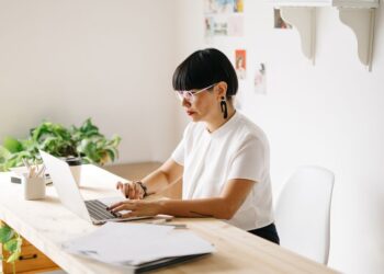 Stylish adult woman using laptop at wooden table in contemporary trendy studio working in daylight