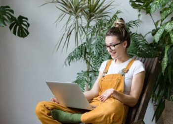 Woman gardener in glasses wearing overalls, sitting on wooden chair in greenhouse, using laptop after work, talks with her friend about coronavirus and stay home during online video call. Working home