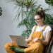 Woman gardener in glasses wearing overalls, sitting on wooden chair in greenhouse, using laptop after work, talks with her friend about coronavirus and stay home during online video call. Working home