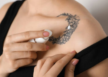 Woman applying cream onto tattoo on her skin against light background, closeup